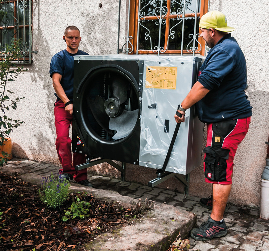 Heizungsmonteure stellen das Außengerät einer Wärmepumpe auf die Wandhalterung.