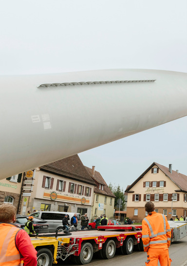 Arbeiter steuern ein großes, auf einem Anhänger transportiertes Windradblatt durch eine Kleinstadtstraße. - © Foto: Uhl Windkraft