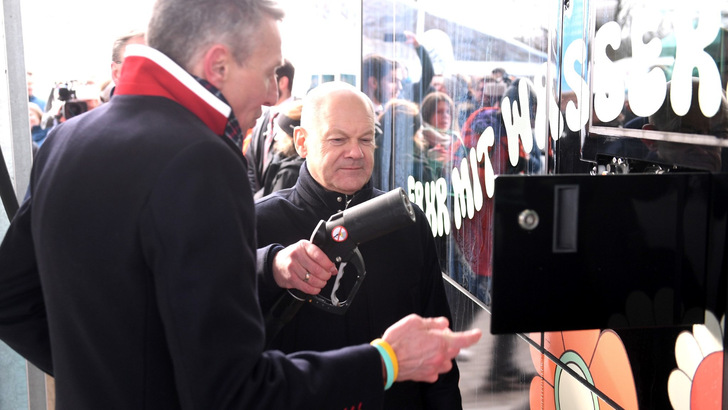 Bei einer öffentlichen Veranstaltung interagieren zwei Männer mit einem Display mit großem Text und reflektierender Oberfläche. - © Energiequelle