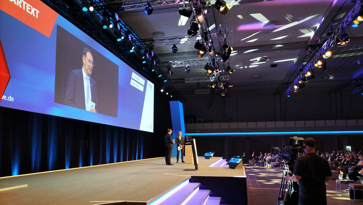 VKU-Verbandstagung 2025 in Berlin: VKU-Hauptgeschäftsführer Ingbert Liebing erklärt die Stadtwerkeforderung nach einem „Neustart der Energiewende“. VKU-Präsident Ulf Kämpfer (hinten), Kieler Oberbürgermeister, verweist darauf, dass der „Kanzler in spe“ zur selben Zeit gerade das „Grünen verstehen“ übe.    