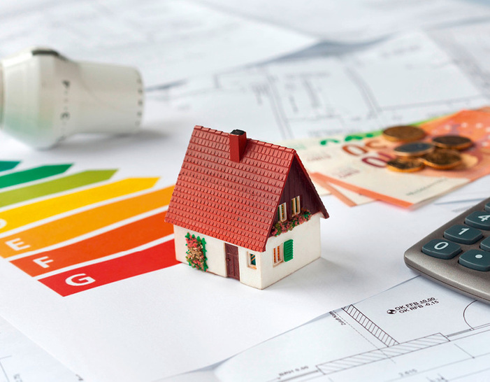 Ein Modellhaus auf Energieeffizienzdiagrammen, mit Euroscheinen, Münzen und einem Taschenrechner in der Nähe. - © Bild: USeePhoto - stock.adobe.com