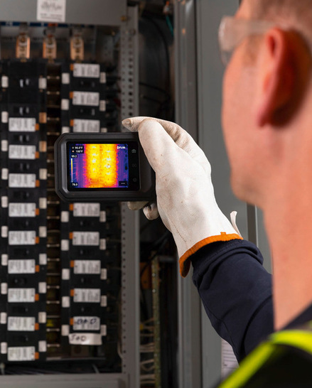 Eine Person verwendet eine Wärmebildkamera, um eine elektrische Schalttafel auf heiße Stellen zu untersuchen. - © Bild: Flir