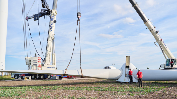 Zwei Kräne heben ein Windradblatt an, während zwei Arbeiter mit roten Helmen die Installation im Freien überwachen. - © HAGEDORN