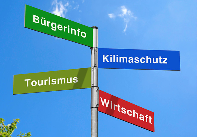 Vier Straßenschilder auf Deutsch an einem Mast: Bürgerinfo, Klimaschutz, Tourismus, Wirtschaft, vor blauem Himmel. - © Foto: hkama - stock.adobe.com