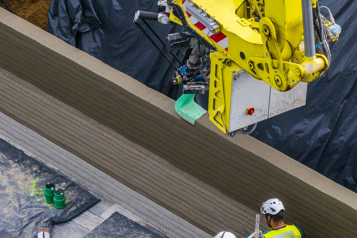 Der Drucker verbindet bewährte Baumaschinentechnik mit Robotik und setzt digitale Entwurfsdaten direkt auf der Baustelle um. Nach Angaben der Projektpartner reduziert die Technologie den Materialverbrauch auf das tatsächlich Erforderliche und senkt den CO₂-Ausstoß gegenüber konventionellem Mauerwerk deutlich.