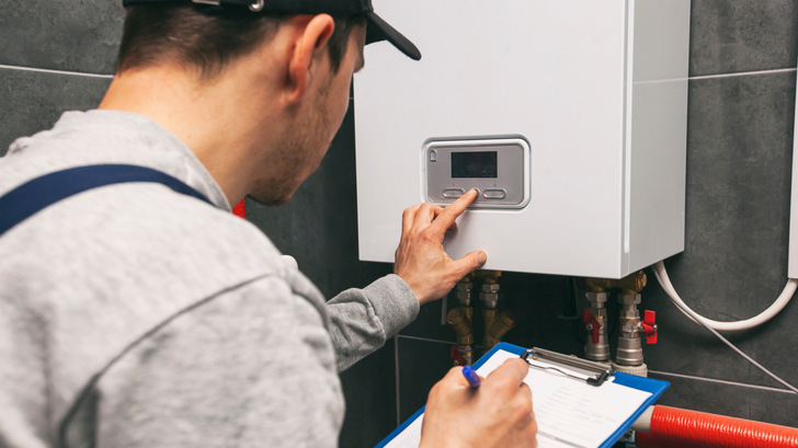 Technician servicing holding clipboard and inspecting heating system in boiler room