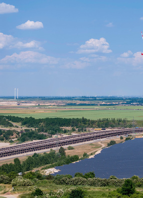 Eine Landschaft mit einem Fluss, grünen Feldern, einem Förderband und Windrädern unter einem blauen Himmel mit Wolken. - © Foto: RWE