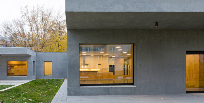 Modernes graues Betongebäude mit großen Fenstern und warmer Innenbeleuchtung, umgeben von Gras und Bäumen. - © Bild: Alexander Blumh﻿off